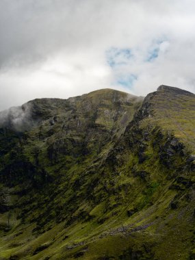 Yeşil çimenler ve bulutlarla kaplı kayalık Coomloughra At Nalı Dağları manzarası.