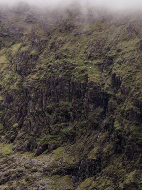 Yeşil bitki örtüsüyle kaplı dik ve sarp bir dağ yamacı keskin bir şekilde yükselir. Sis zirveyi gizliyor. Bu dağ İrlanda 'nın Kerry ilçesinde..