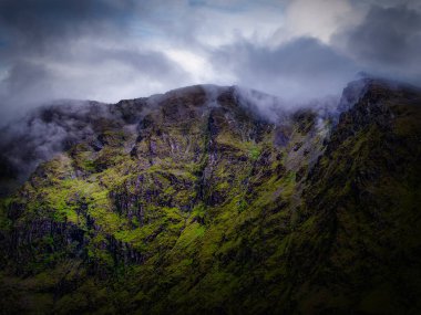 Dik dağ yamacı kısmen alçak bulutlarla gizlenmiş dramatik bir manzara. Yeşil bitki örtüsü yumuşak, doğal ışık altında kayalara tutunur..