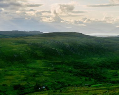 Yuvarlanan yeşil tepeler bulutlu bir gökyüzünün altındaki manzarayı kaplıyor. Dağlar uzaktan görülebilir. Ağaçların arasına küçük bir bina kurulmuş..