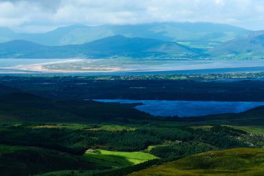 Yeşil tepeler ve ormanlar mavi bir göl ile buluşur. Dağlar parlak, bulutlu bir ufukta kaybolur. Yeşil katmanlı uçsuz bucaksız bir arazi.