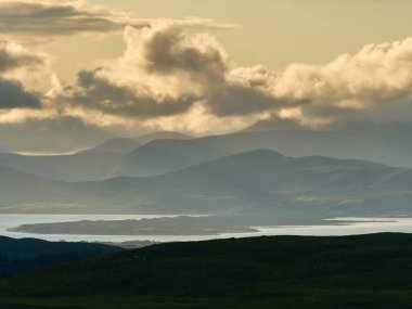 Ufuk boyunca uzanan katmanlı dağlar Castlemaine Limanı 'nın sakin sularını çerçeveliyor. Altın ışık, dramatik bulutların arasından süzülür, sakin manzaranın üzerine yumuşak tonlar saçar..