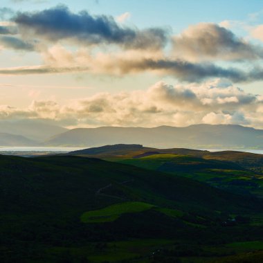 Yuvarlanan yeşil tepeler dramatik bir gökyüzü altında puslu dağlarla Castlemaine Limanı 'na çıkar. Altın ışık, sakin bir akşamda bulutları ve manzaranın bazı kısımlarını aydınlatır..