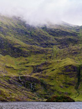 Görkemli yeşil bir dağ, kısmen sisle kaplı, yamaçlarından aşağı doğru akan birkaç dar şelaleyle birlikte sakin bir göle hakim. Suda küçük bir ada var..