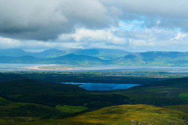 Yeşil tepeler ve ormanlar mavi bir göl ile buluşur. Ormanlar, su ve dağlarla kaplı bir manzara..