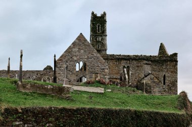 Timoleague, İrlanda, 25 Kasım 2023. Bir ortaçağ manastırının taş kalıntıları ve birçok haçı olan eski bir mezarlık, gri, bulutlu İrlanda kırsalındaki bir gökyüzünün altında yeşil, çimenli bir tepenin üzerinde yer almaktadır..