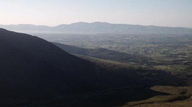 Uçsuz bucaksız kırsal bir vadide yumuşak akşam ışığı ve yüksek bir tepenin gölgeli yamacından görünen uzak puslu dağlar..