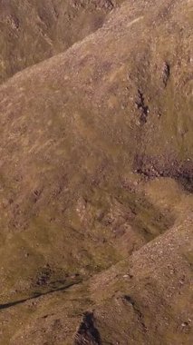 Parlak gün ışığında yıkanan kurak bir dağ sırasının geniş bir görüntüsü. Dik, kayalık tepeler ve vadiler görülebilir, uzun gölgeler manzaranın dramatik özelliklerini ve düz olmayan yüzeyini vurgular..