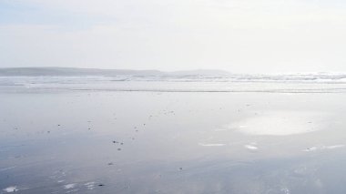 Geniş, ıslak kumlu bir sahil, parlak ve puslu bir gökyüzünün altında okyanusa doğru uzanır. Yumuşak dalgalar uzaktaki ince kıyı şeridi boyunca kırılır..