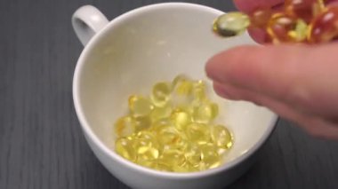 Bir avuç dolusu Omega 3 balık yağı kapsülü avucunun içinden ahşap bir arka planda beyaz bir bardağa düşer. Yakın plan. Yavaş çekim