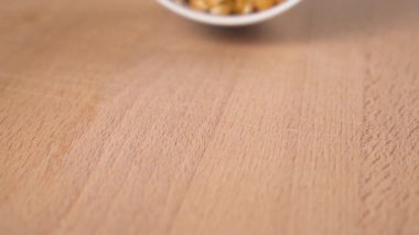 Kurumuş mısır tohumları beyaz bir bardaktan yavaş çekimde tahta bir yüzeye düşer. Kapatın. Patlamış mısır yapmaya hazırlanıyor.