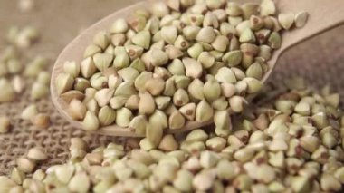 Biyolojik ürün. Yeşil, tahta bir kaşıkta çuvalın üstünde pişmemiş karabuğday. Düşen çiğ karabuğday taneleri. Çuval bezi dokusu. Makro. Yavaş çekim