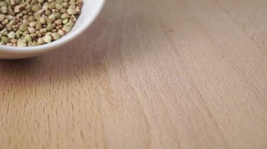 Biyolojik ürün yeşil karabuğday yavaş çekimde beyaz bir kasenin ahşap tahtasına saçılmıştır. Yakın plan.