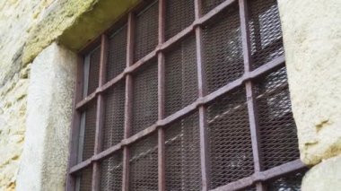 Antika bir binanın eski taş duvarında paslı ızgarası ve demir örgüsü olan parmaklıklı eski bir pencere. Pencerenin camında yansıma var. Dramatik Görünüm