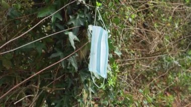 Kırsal kesimde bir tel örgüde kullanılmış bir maske, ahşap ve bitki örtüsüne karşı. Rüzgarda savruluyor. Koronavirüs tehlikesi ve ekoloji kirliliği kavramı