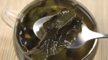 Çay kaşığında demlenmiş yeşil çay yaprağı bir fincan sıcak aromatik içeceğin üzerinde. Bulanık bir bambu hasırın üzerinde. Doğal bitki çayı demliyorum.