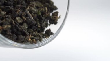 Kurutulmuş yeşil çay yaprakları cam bir fincanın içinde. Beyaz bir yüzey üzerinde demlenmemiş çayı yavaş çekimde dökülüyor.