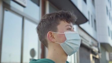 Tıbbi tek kullanımlık koruyucu maskeli neşeli ve yakışıklı bir gencin portresi bulanık bir şehir üniversitesi binasının arka planında. Öğrenci başını çevirir ve güler.