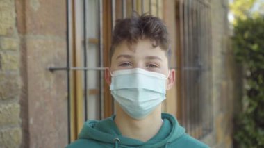 Tıbbi koruyucu maskeli esmer ve gür saçlı genç bir adam binanın kafes pencereleri ve bulanık bitki örtüsünün arka planına gülüyor. Yakın plan portre. Karantina ve tecrit kavramı