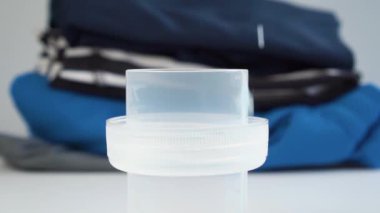 Plastik kapak yavaş çekimde mavi sıvı çamaşır deterjanıyla doludur, arkasında da bulanık renkli giysiler vardır. Kapatın. Ev aletleri konsepti