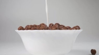 Süt, çikolatalı gevrek toplarıyla dolu beyaz bir kaseye dökülüyor. Ağır çekim. Ön görünüm