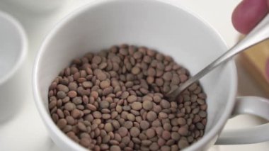 Mutfak masasının üzerinde beyaz bir bardakta kaşıkla çiğ mercimek karıştırmak. Ağır çekim. Geleneksel yemekler pişirmek