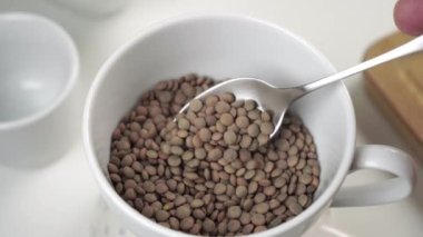 Mutfak masasındaki beyaz bardakta pişmemiş kahverengi mercimek. Metal kaşıkla karıştır. Makro çekim