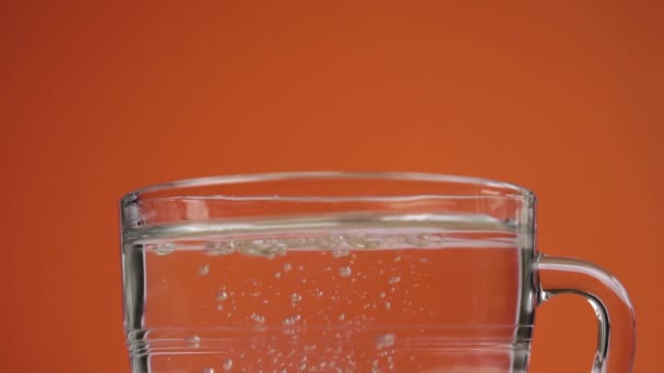 効果的な可溶性錠剤は スプラッシュときれいな飲料水のガラスカップに落ち 液体の表面に浮かぶ泡で溶解します スローモーション 閉めろ 鮮やかなオレンジ色のテラコッタの背景に — ストック動画
