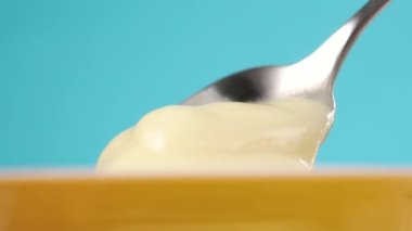 Tatlı kaşığı, açık sarı plastik bir kaptan tatlı kremalı yoğurt topluyor. Süt ürünü konsepti. Renkli mavi arka planda çırpılmış ekşi süt. Makro çekim