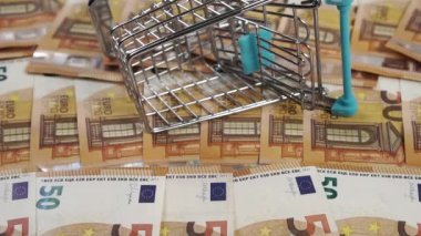 Bir tomar Avro parası, arka planda Avrupa banknotları istiflenmiş tüketici mini alışveriş arabasının yanına düşüyor. Ağır çekim. Yakın plan. Zenginlik ve alışveriş kavramı