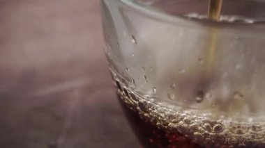 Ağır çekimde, buhar ve kabarcıklarla cam fincana koyu siyah çay dökülür. Makro. Bulanık kahverengi bir tezgahın üzerinde.