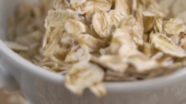 Bir kaşık, ağır çekimde beyaz bir kaseden pişmemiş yulaf ezmesi topluyor. Makro. Dokulu yulaf lapası malzemesi. Çok yakın çekim.