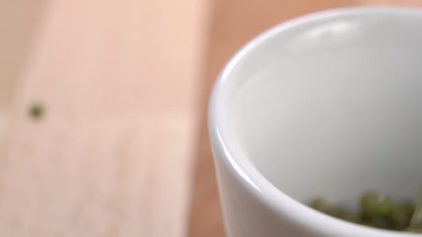 生の緑の豆は白いカップを満たし ぼかしの遅い動きで木製の表面全体に散布します マクロショット 健康的なアジア料理のコンセプト — ストック動画