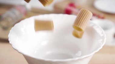 Pişmemiş rigatoni integral makarna yavaş çekimde mutfak masasının üzerindeki beyaz bir kaseye düşer. Yakın plan.