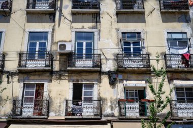 A close-up of a typical, sun-drenched, run-down yellow facade in Lisbon, featuring old white windows, ornate black wrought iron balconies, and utility cables