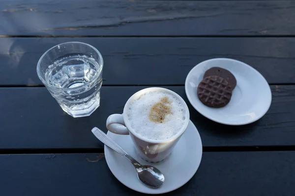 Cup of milk with coffee, sparkling water and a chocolate cookie. On a black table.