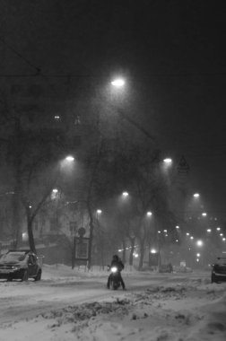 Bisikletli teslimatçı. Karlı bir akşam caddesinde ilerliyor. Siyah beyaz.