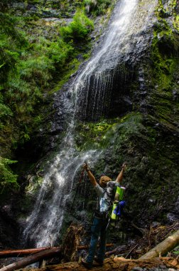 Yeşil şelalede poz veren sırt çantalı bir yürüyüşçü.