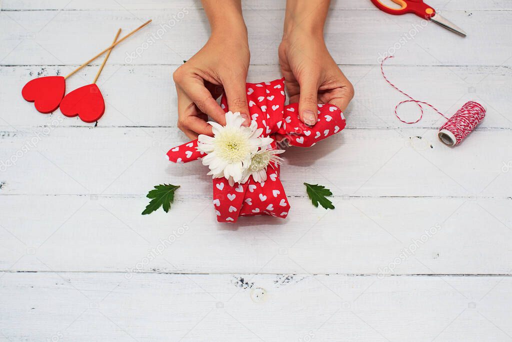 Womens hands wrappes gift for Valentine's Day, Birthday, Mother's Day using red cotton textile and flowers on a white wooden background. Furoshiki, ecological gift wrapping