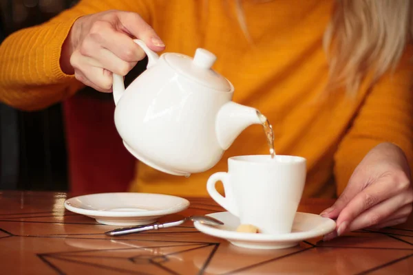 A woman in a yellow sweater pours tea from a white teapot into a cup. Tea drinking in a cafe or at home.