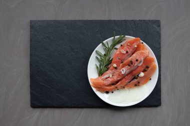 Baharatlı deniz alabalığı parçaları ve siyah bazalt tahtasındaki beyaz bir porselen tabakta duran biberiye filizi.