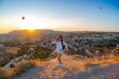 Sırt çantalı bir kız gün doğumunda dağ ve balonlarla vadide poz veriyor. Eğlence, turizm bir tatil. Seyahat turu. Goreme, Kapadokya, Türkiye.