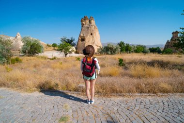 Gezgin kız sırt çantalı, deri şapkalı, turist pozu veren. Arka planda peri bacaları var. Kaya oluşumları. Seyahat ve tatil, tur Goreme, Kapadokya, Türkiye.