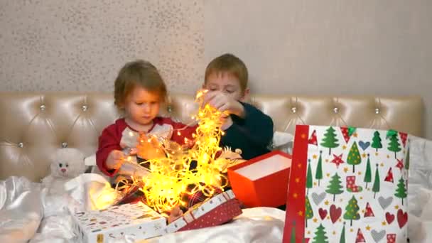 Kinderen zitten op het bed, halen een kerstslinger uit een mooie doos en spelen ermee. Ze kijken naar de lichten en ontwarren de draden. Ze hangen een bloemenslinger aan hun lichaam. — Stockvideo