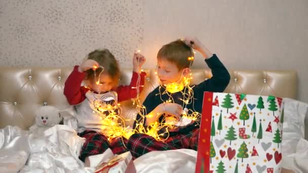孩子们坐在床上，从一个漂亮的盒子里拿出一个圣诞花环来玩。他们看着灯，解开电线。他们把一个花环挂在身上 — 图库视频影像
