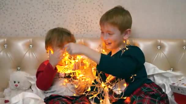 Kinderen zitten op het bed, halen een kerstslinger uit een mooie doos en spelen ermee. Ze kijken naar de lichten en ontwarren de draden. Ze hangen een bloemenslinger aan hun lichaam. — Stockvideo