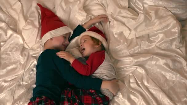Brat i młodsza siostra leżą na łóżku, przytulają się i śmieją radośnie i żarliwie. są ubrane w piżamy i kapelusze noworoczne — Wideo stockowe