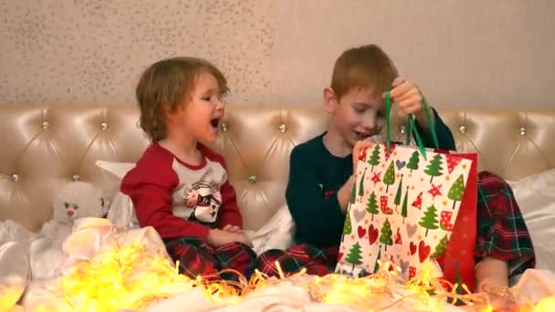 As crianças sentam-se na cama e tiram surpresas. Do pacote, eles tiram uma lanterna-uma luz noturna em que Papai Noel com uma árvore de Natal e brilho voar como flocos de neve em uma tempestade de neve — Vídeo de Stock