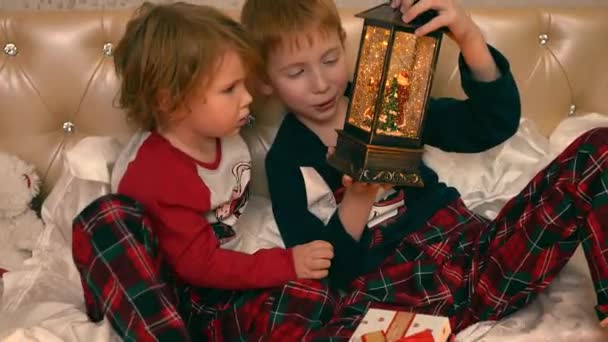 Niños se sientan en la cama y miran la luz de la linterna de la noche, en la que Papá Noel con un árbol de Navidad y lentejuelas vuelan como copos de nieve en una tormenta de nieve — Vídeos de Stock