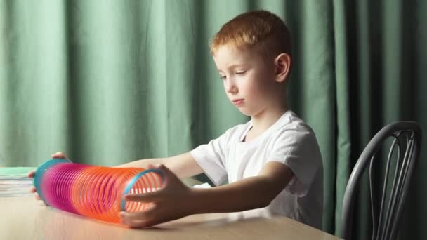 Le garçon roux assis à la table est fatigué de faire les devoirs de l'école et il joue avec un jouet printemps slinky. Il a mis ses livres et son carnet. — Video